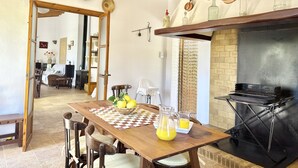 A dining area with a wooden table, chairs, and a pitcher of juice. There is a brick fireplace and a grill in the background. - Es Carritxó, Peace and Nature in Mallorca (Carritxo (Es))
