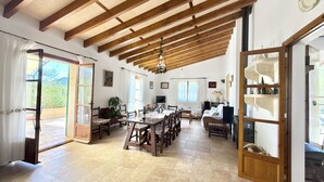 A spacious dining area with a high ceiling and wooden beams, a large table with chairs, and a view of the outdoors through glass doors. - Es Carritxó, Peace and Nature in Mallorca (Carritxo (Es))