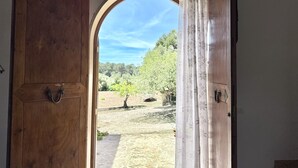 An open wooden door leading to an outdoor area with a stone pathway, a tree, and a view of a green landscape under a clear sky. - Es Carritxó, Peace and Nature in Mallorca (Carritxo (Es))