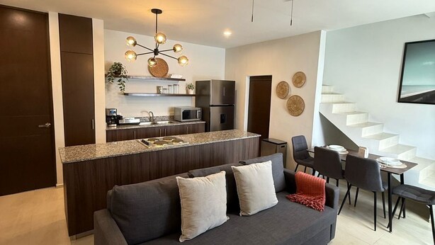 A modern living room with a dark grey sofa, a ceiling fan, a kitchen area with a stainless steel refrigerator, and a staircase leading to an upper level. - Apartment with Private Rooftop. Start your adventure here at Zefira - 2BR. (Mérida)