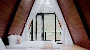 A bedroom with a sloped ceiling, a bed with white bedding, and a view through a large window. - Dieng Ceria By Travelink (Dieng Kulon)