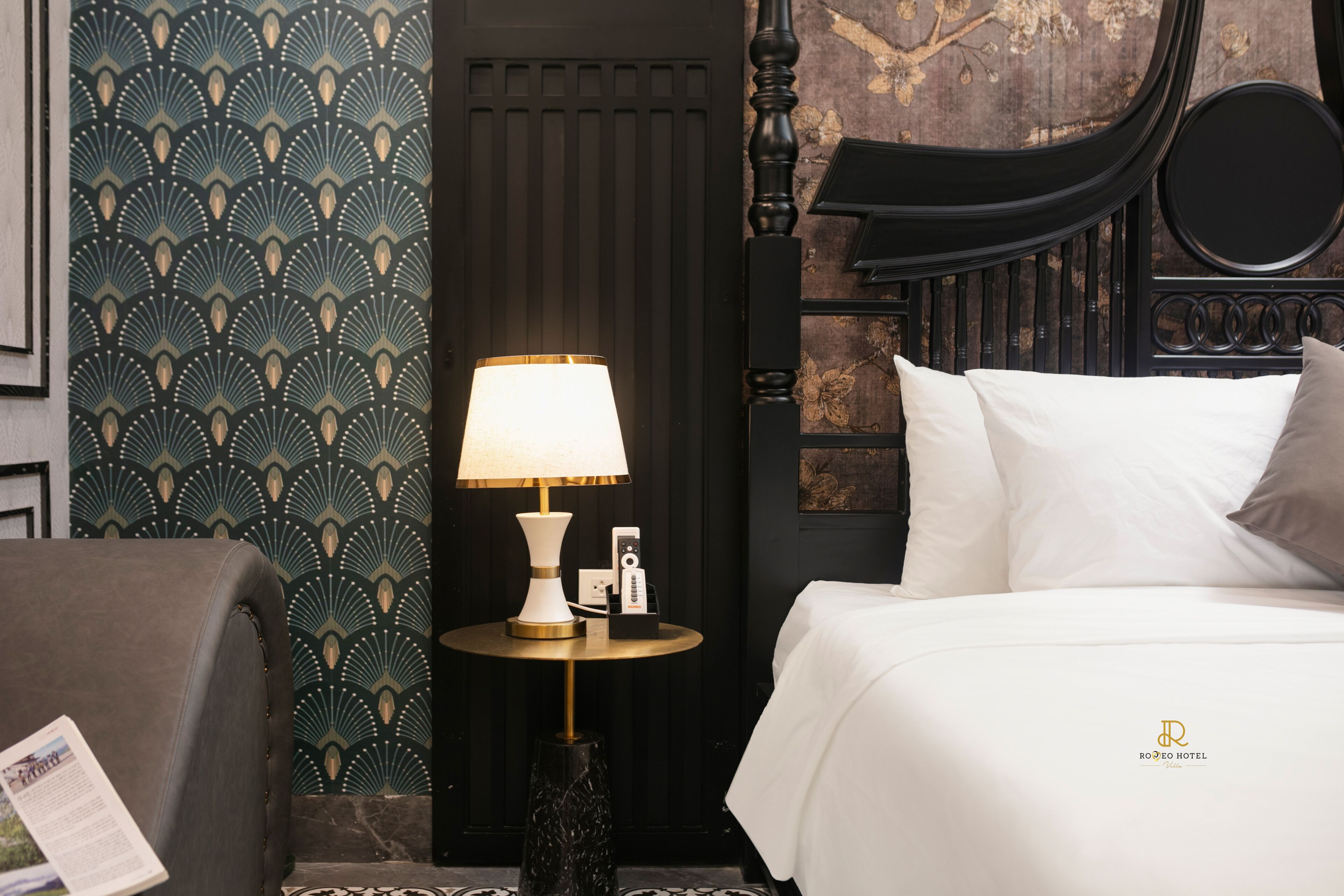 A hotel room with a bed, a round table with a lamp, a chair, and a patterned wallpaper.