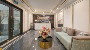 A hotel lobby with a reception desk, a sofa, a coffee table with flowers, and a large mirror. - Romeo Hotel Linh Dam 1 - by BAY LUXURY (Hanoi)