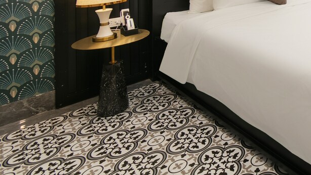 A hotel room with a bed, a nightstand with a lamp, a decorative wall, and patterned flooring. - Romeo Hotel Linh Dam 1 - by BAY LUXURY (Hanoi)