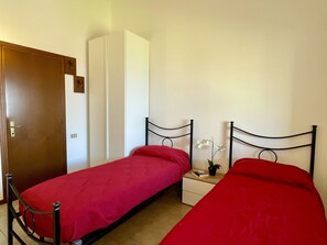 Un petit logement composé d’une seule pièce, comprenant deux lits simples, une armoire, une table de chevet avec une plante et une porte.