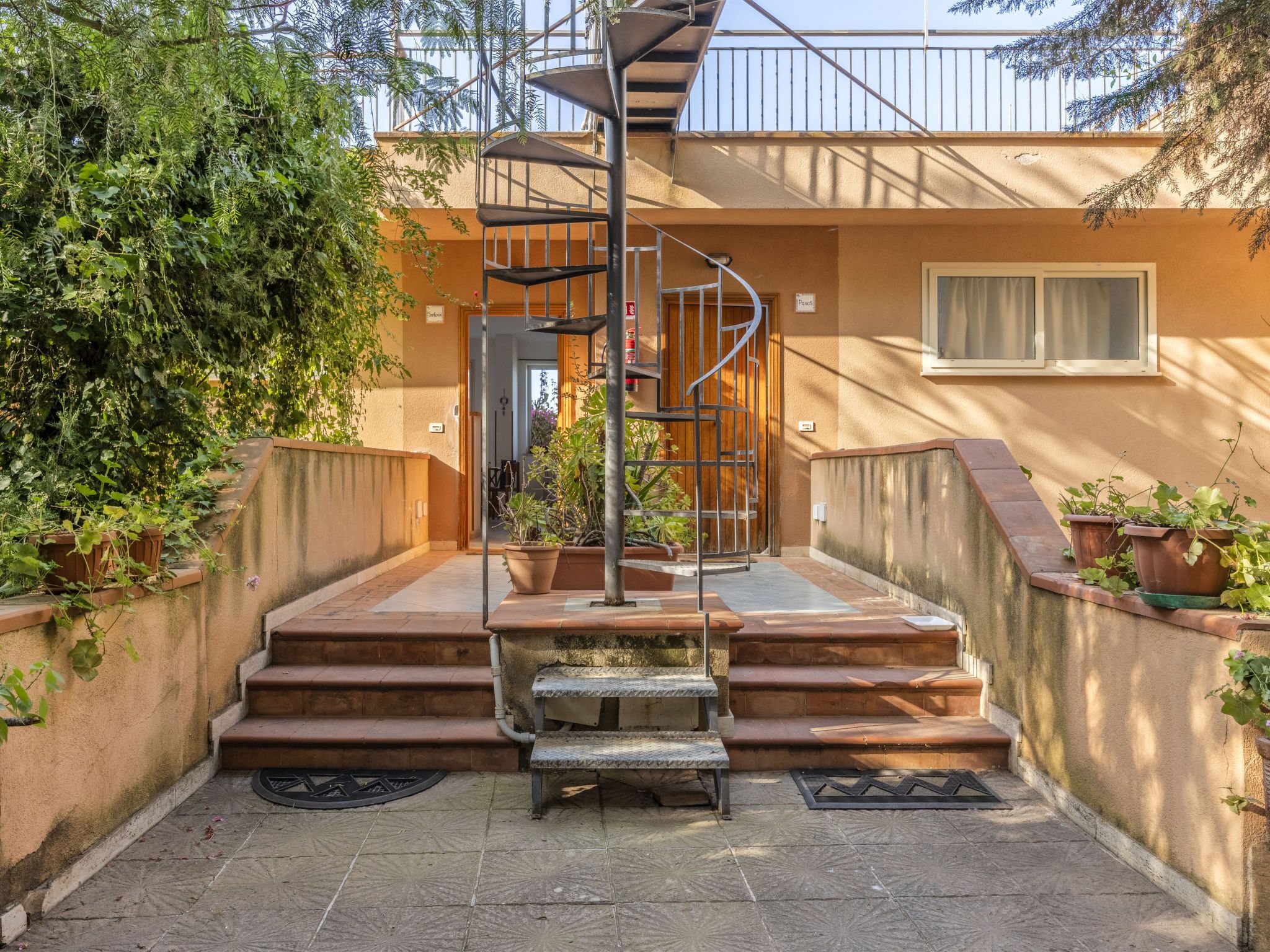 A spiral staircase leading to a door, surrounded by potted plants and a tiled patio.
