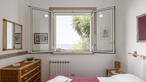 Un dormitorio con cama, cómoda, ventana con contraventanas abiertas y radiador.
