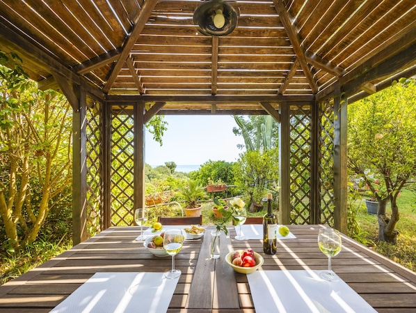 A wooden pergola with a table set for dining, surrounded by lush greenery and a view of the ocean.