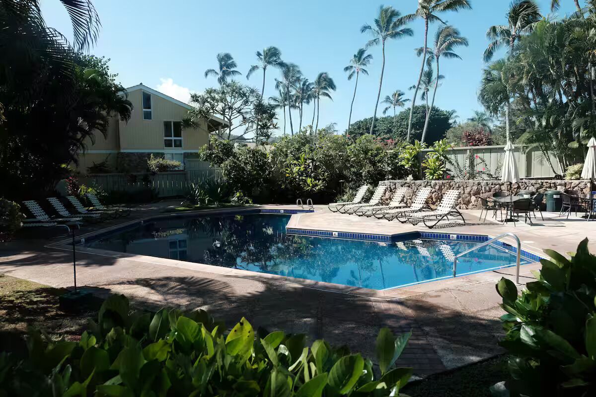 A swimming pool with lounge chairs and umbrellas, surrounded by palm trees and a building.
