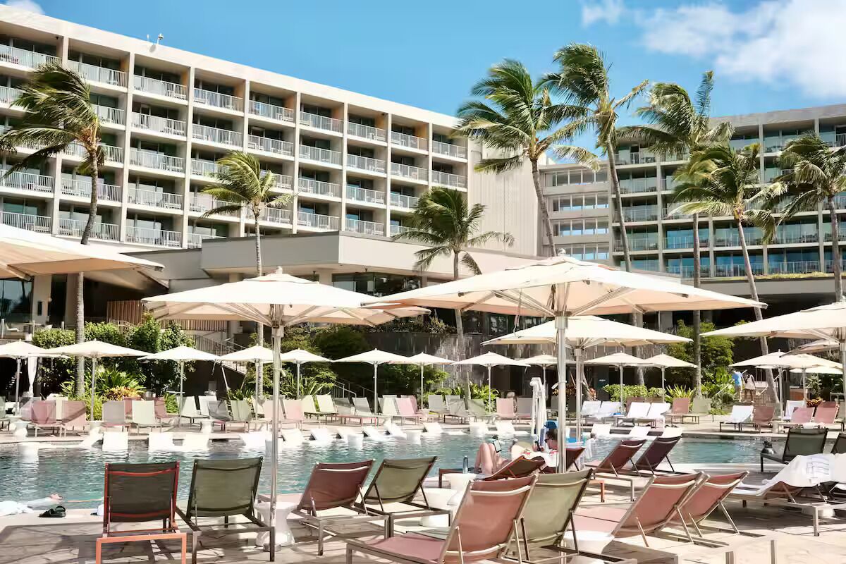 Een zwembadgedeelte van een hotel met meerdere ligbedden en parasols, omgeven door palmbomen en een meerlaags gebouw met balkons.
