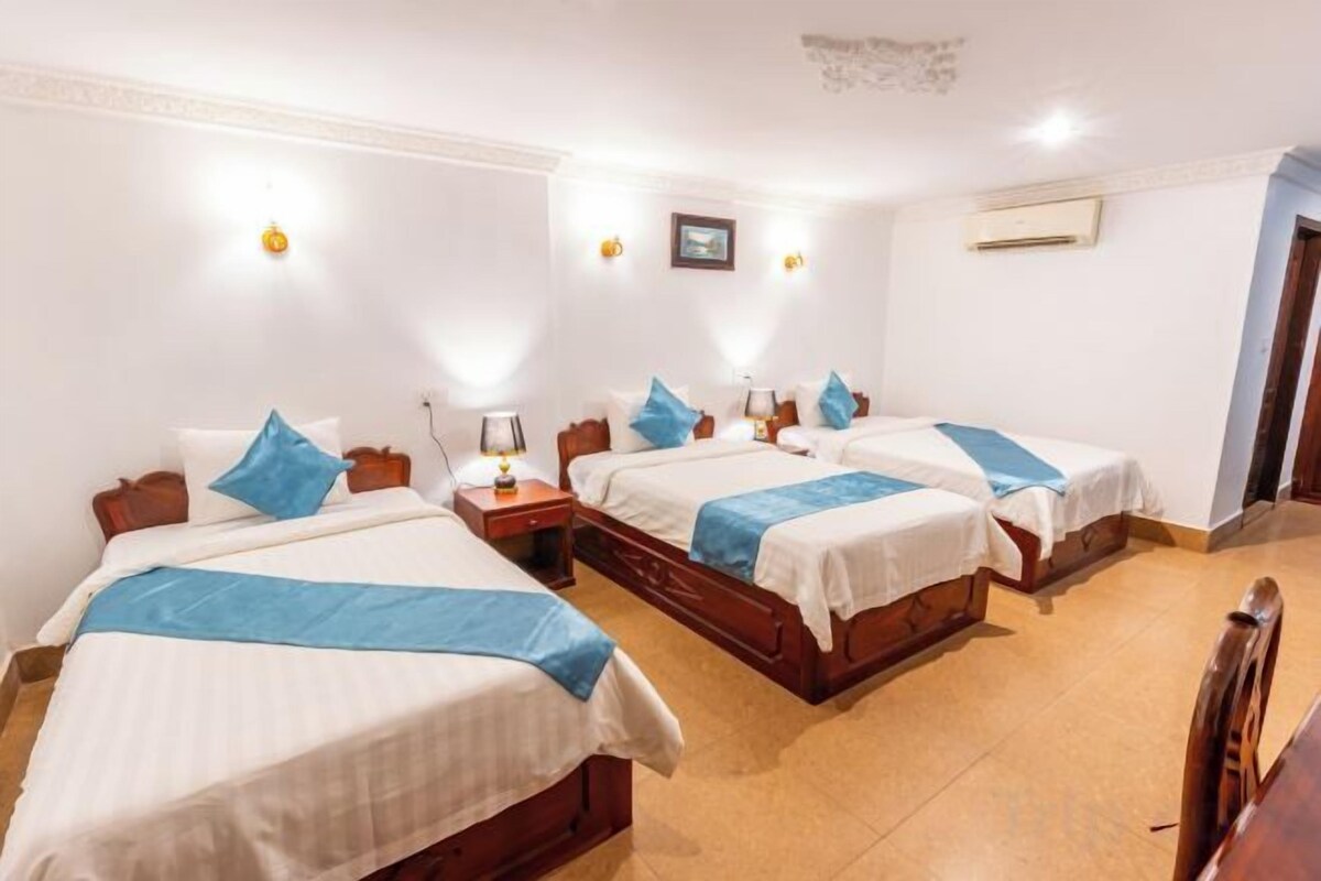 A hotel room with four single beds, each with a blue accent pillow and a white bedspread. There are bedside tables with lamps, and the room has a tiled floor.