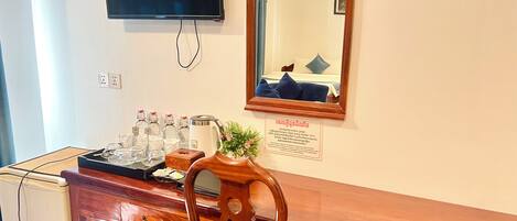 A hotel room with a wooden desk, a television mounted on the wall, a mirror, and a sign with Thai text.