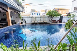 A swimming pool with lounge chairs and a covered seating area, surrounded by buildings and greenery. - Rithy Rin Angkor Hotel (Siem Reap)