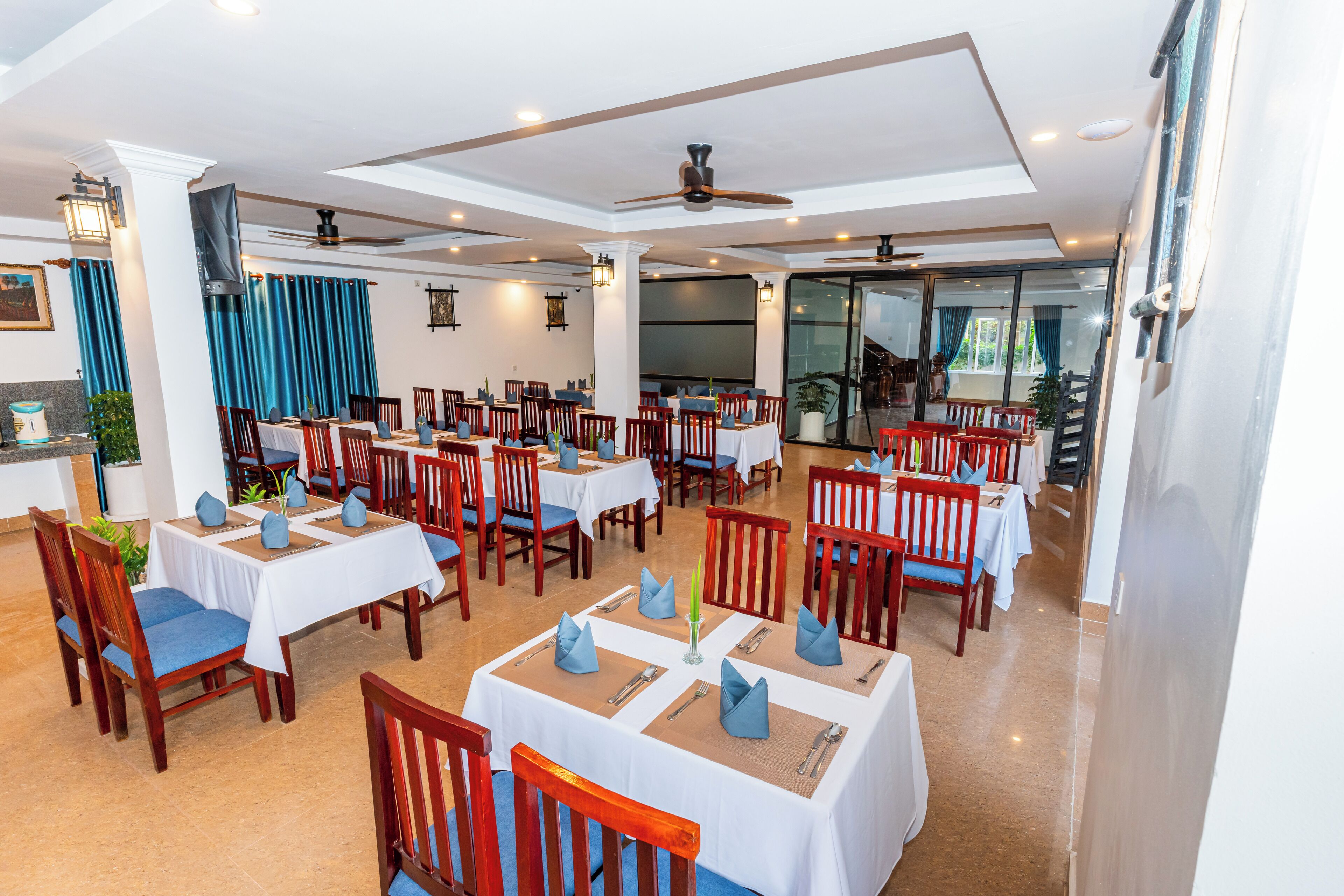 A restaurant with multiple tables set with blue napkins and white tablecloths, wooden chairs, and ceiling fans.
