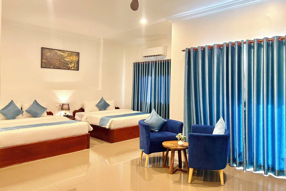 A hotel room with two beds, a ceiling fan, a small table with a vase, and blue curtains.