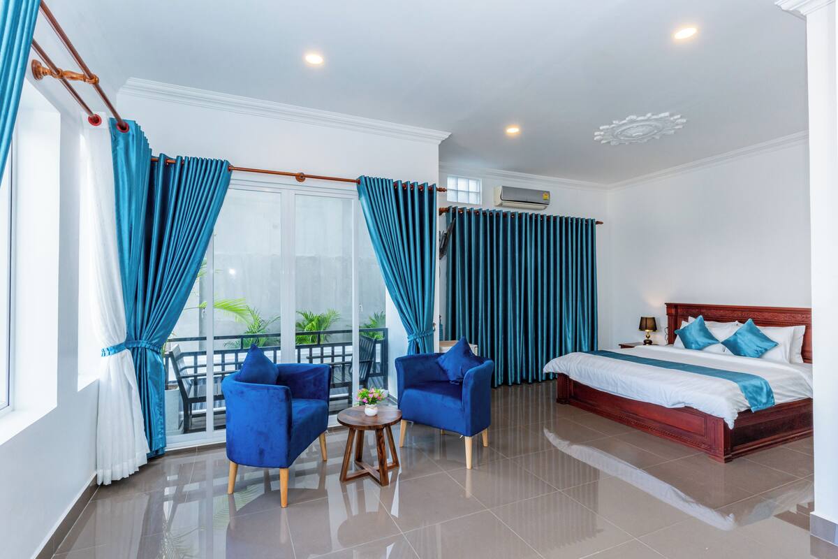 A hotel room with a bed, two blue armchairs, a small table with a vase, and a balcony with blue curtains.