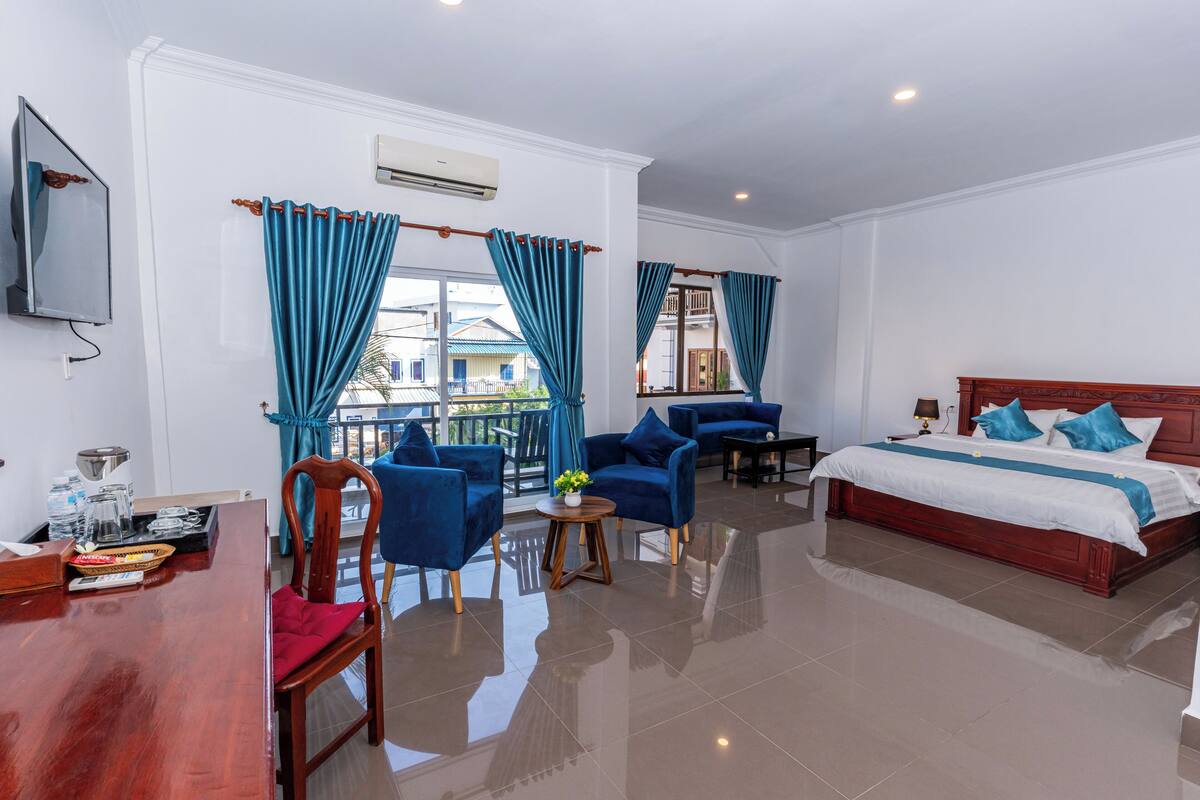 A hotel room with a bed, two blue armchairs, a wooden table, a TV, and a balcony with blue curtains.