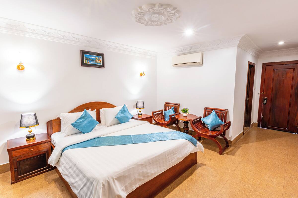 A hotel room with a large bed, a wooden nightstand, a table with chairs, and a door.
