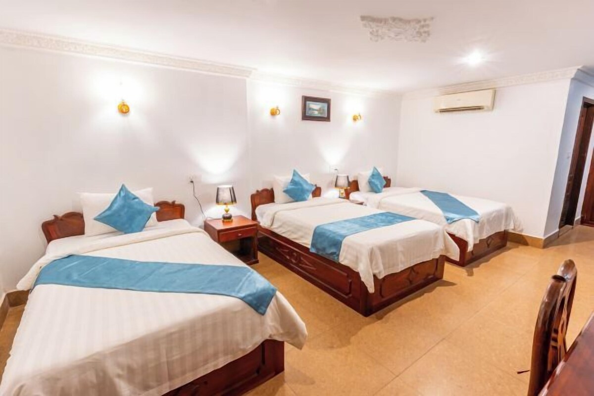 Une chambre d’hôtel avec trois lits simples, chacun agrémenté d’un coussin bleu et doté de draps blancs. On y trouve des tables de chevet avec des lampes, un climatiseur fixé au mur et un tableau encadré au-dessus des lits.