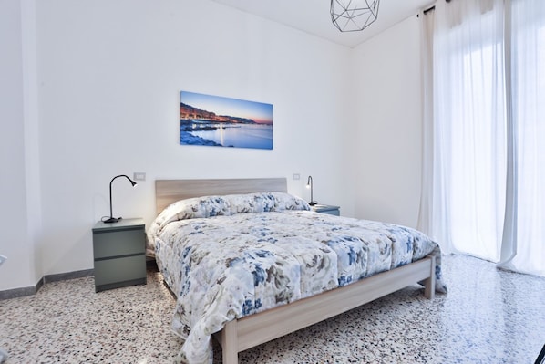 A bedroom with a bed, two bedside tables, a nightstand, a wall-mounted picture, and a window with curtains.