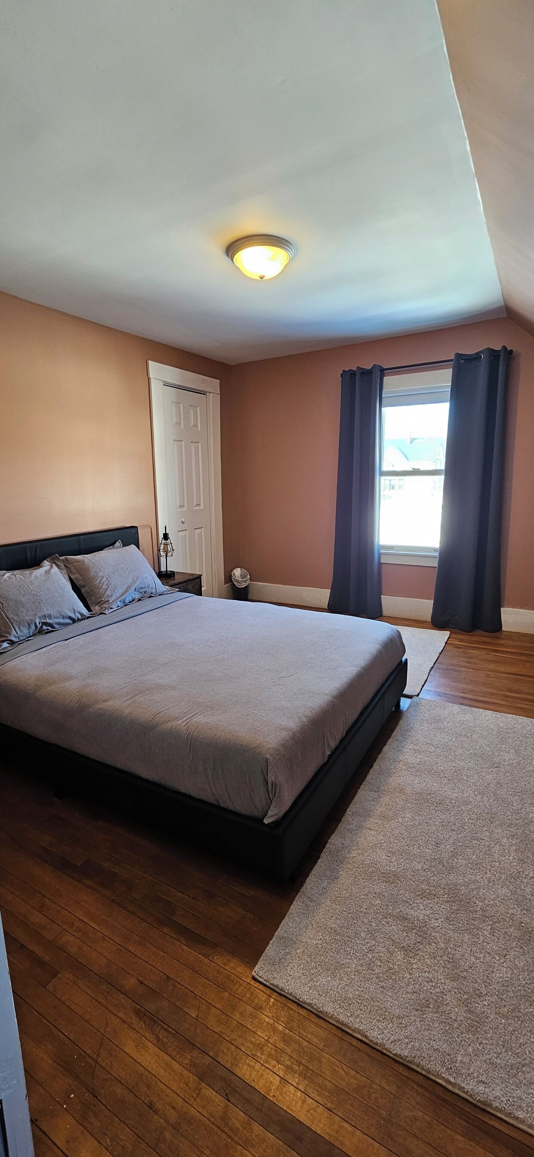 Un dormitorio con cama, mesita de noche, lámpara, puerta y ventana con cortinas.