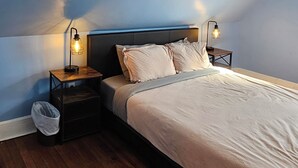 A bedroom with a sloped ceiling, a bed with a beige bedspread, two bedside tables with lamps, a wooden floor, and a small trash bin. - Scenic Shelburne home. Central location close to Shelburne Harbour. (Shelburne)