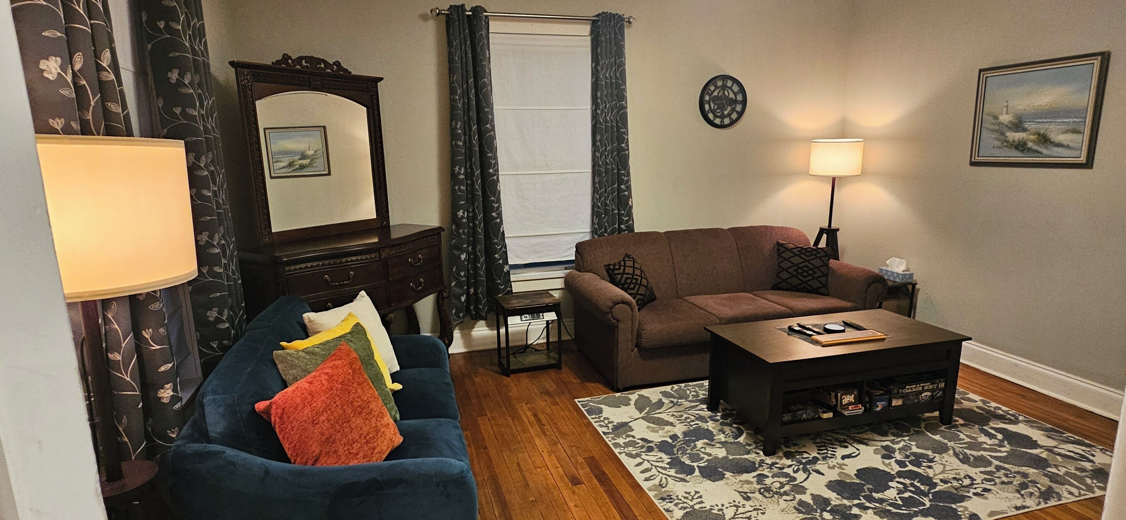 A living room with a sofa, a coffee table, a lamp, a clock, and a painting on the wall.