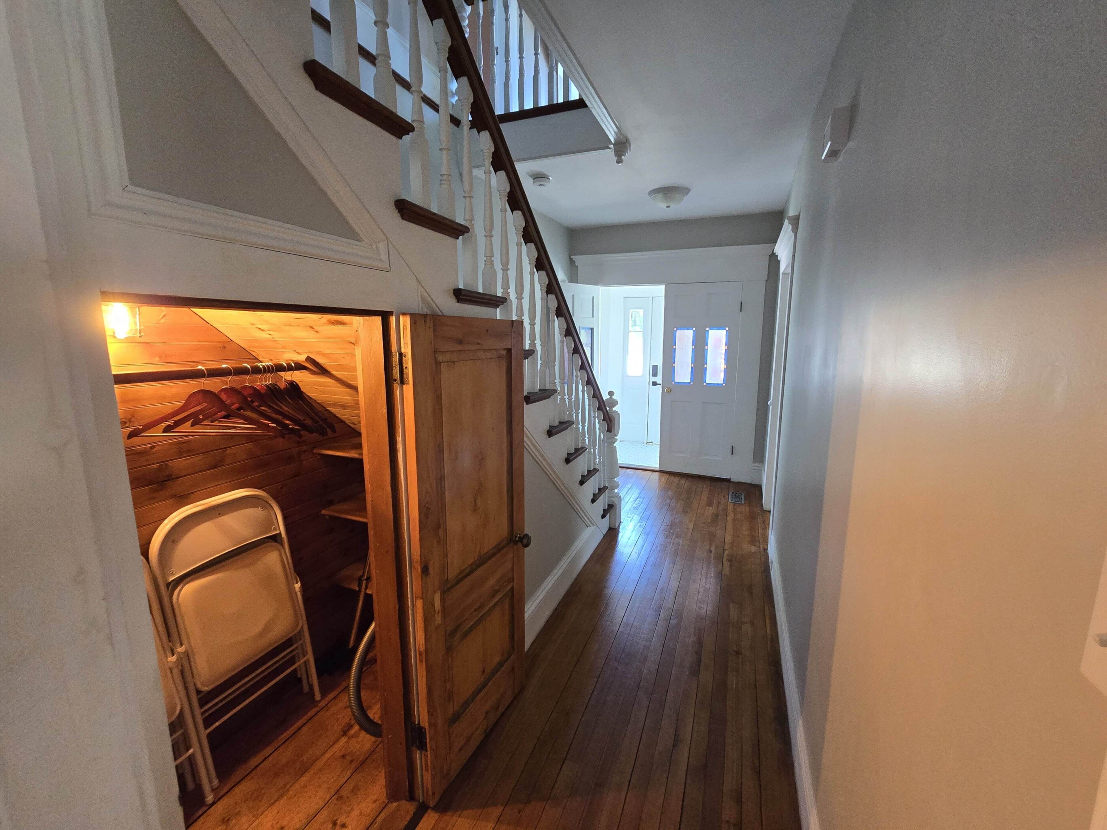 A hallway with wooden flooring, a staircase with white balusters, and a door leading to a room with a wooden closet and a chair.