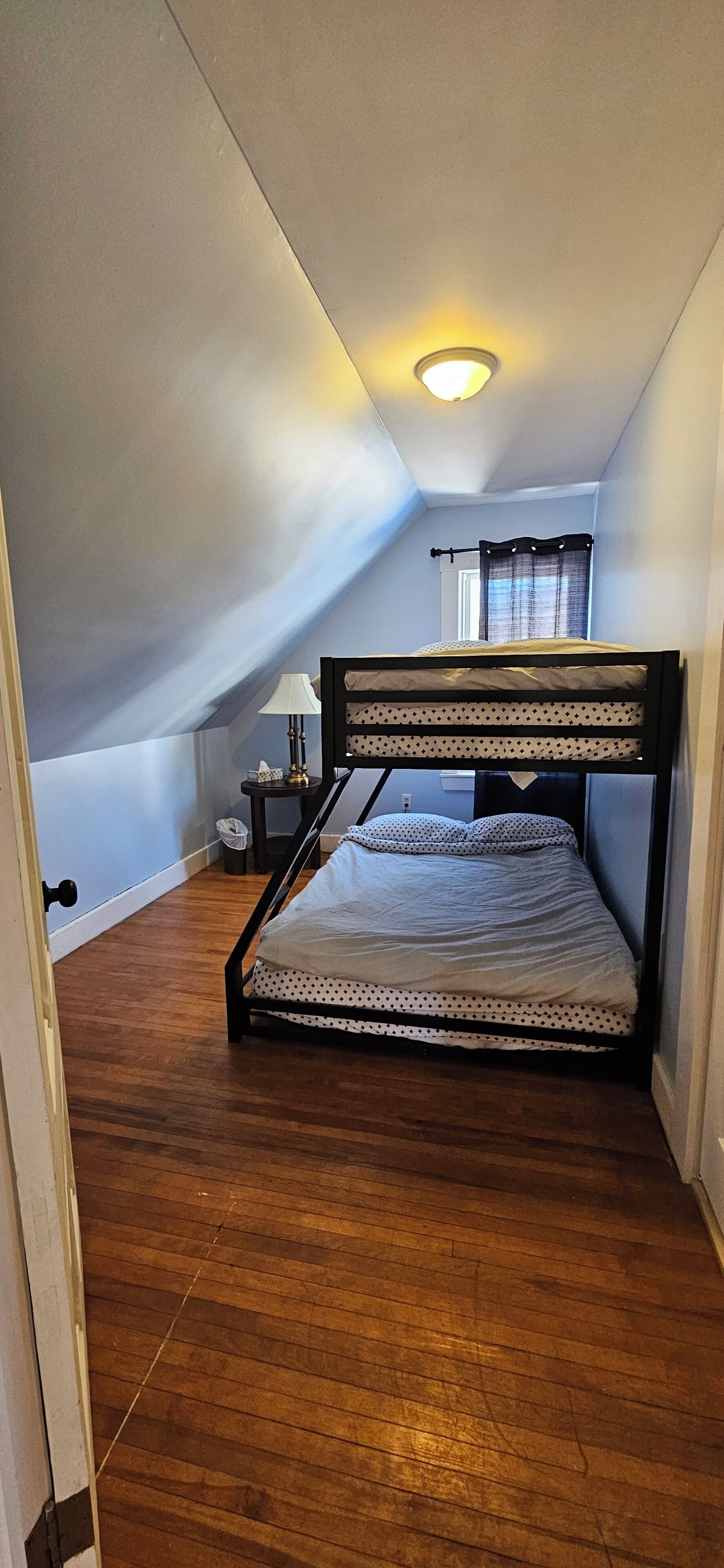 A small, cozy attic bedroom with a bunk bed, wooden flooring, a bedside table with a lamp, and a window with curtains.