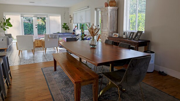 A spacious dining area with a wooden table, benches, and modern chairs. There is a large window with blinds, a cabinet with decorative items, and a view of the outdoors. - Bonita Ranch (Miami Shores)