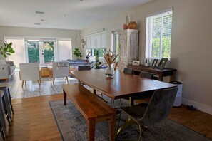 Un'ampia sala da pranzo con tavolo in legno, panche e sedie moderne. Si apre su un'arredamento con tende, una credenza con oggetti decorativi e vista sull'esterno.