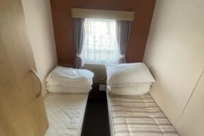 A narrow room with two beds, a window with curtains, and a radiator. - 3 Bed Caravan - Sleeps 8 - Pets - Parking - Wifi (Rhyl)
