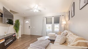 A modern living room with a sofa, a coffee table, and a TV. There is a door leading to another room and a window with curtains. - Quiet Comfort-Parking & Patio • WiFi • WD (Austin)