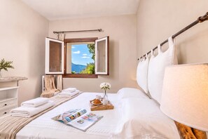 A hotel room with a bed, a window with an open curtain, a bedside table with a lamp, and a view of the outdoors.