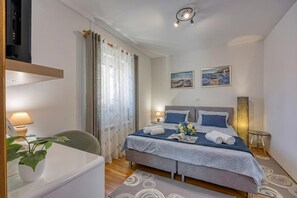 Quarto com cama, mesas de cabeceira, escrivaninha, cadeira, planta em vaso e duas pinturas na parede.