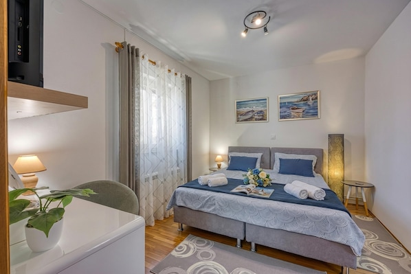 A bedroom with a bed, bedside tables, a desk, a chair, a potted plant, and two paintings on the wall.