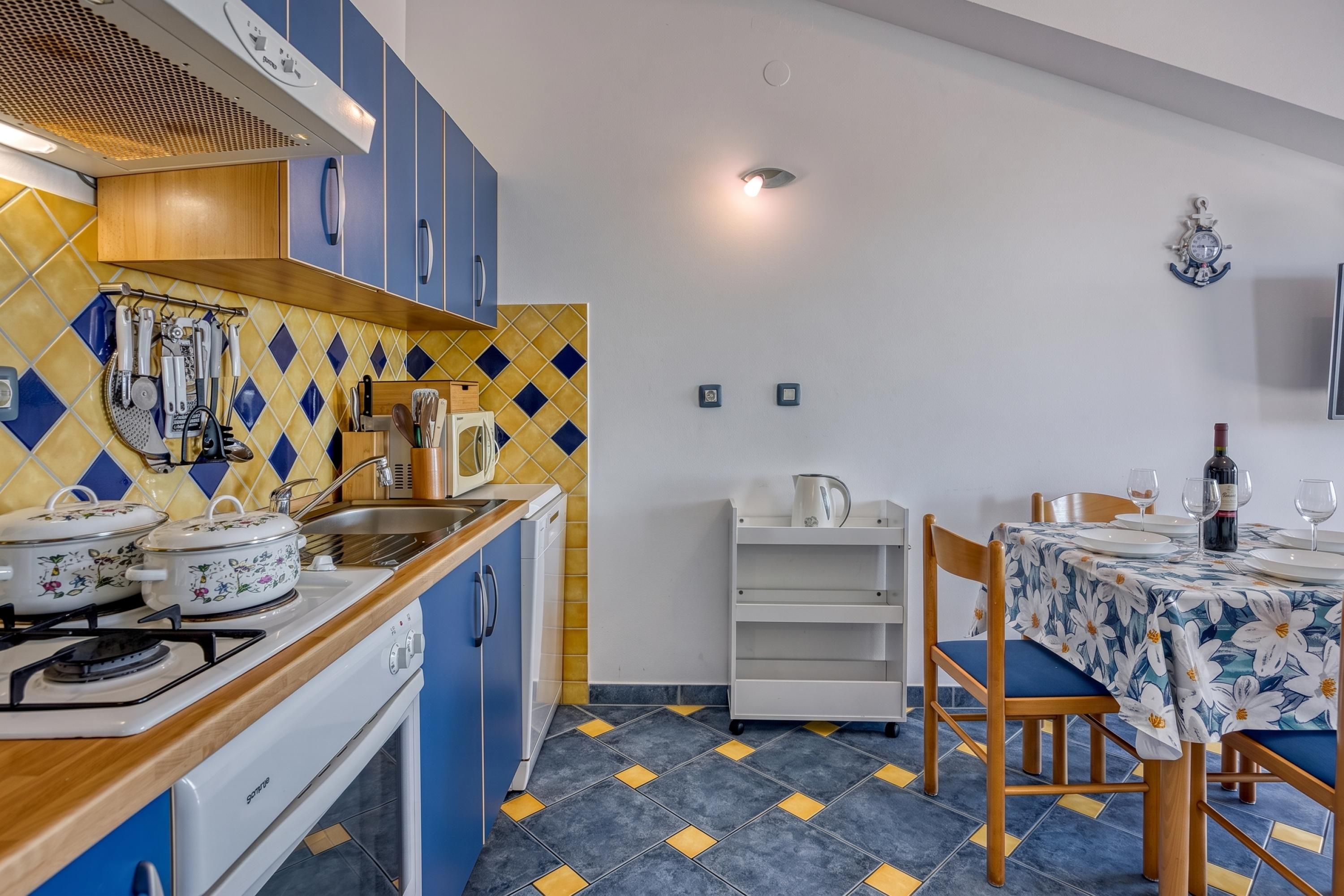 Une cuisine compacte avec des meubles bleus, une cuisinière à gaz et un évier. On y trouve également un coin repas avec une table dressée pour deux personnes et une chaise.