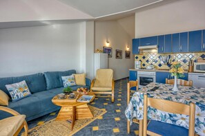 A living room with a sofa, coffee table, and dining table. There is a kitchen with blue cabinets and a microwave.