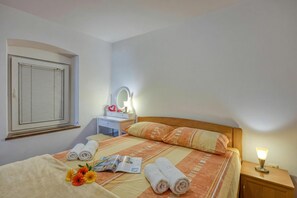 Une chambre d’hôtel comprenant un lit, une table de chevet avec une lampe, une fenêtre avec des stores, un miroir et une petite commode.