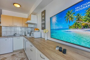 A modern kitchen with white cabinets, a microwave, and a sink. There is a large flat-screen TV on the wall displaying a vacation program.