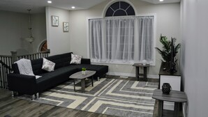 A modern living room with a black sofa, a coffee table, a patterned rug, and a large window with sheer curtains.