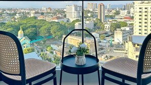 Ein Balkon mit zwei Korbstühlen und einem kleinen Tisch mit Blumenvase, mit Blick auf eine Stadtlandschaft aus Gebäuden und Grünflächen.