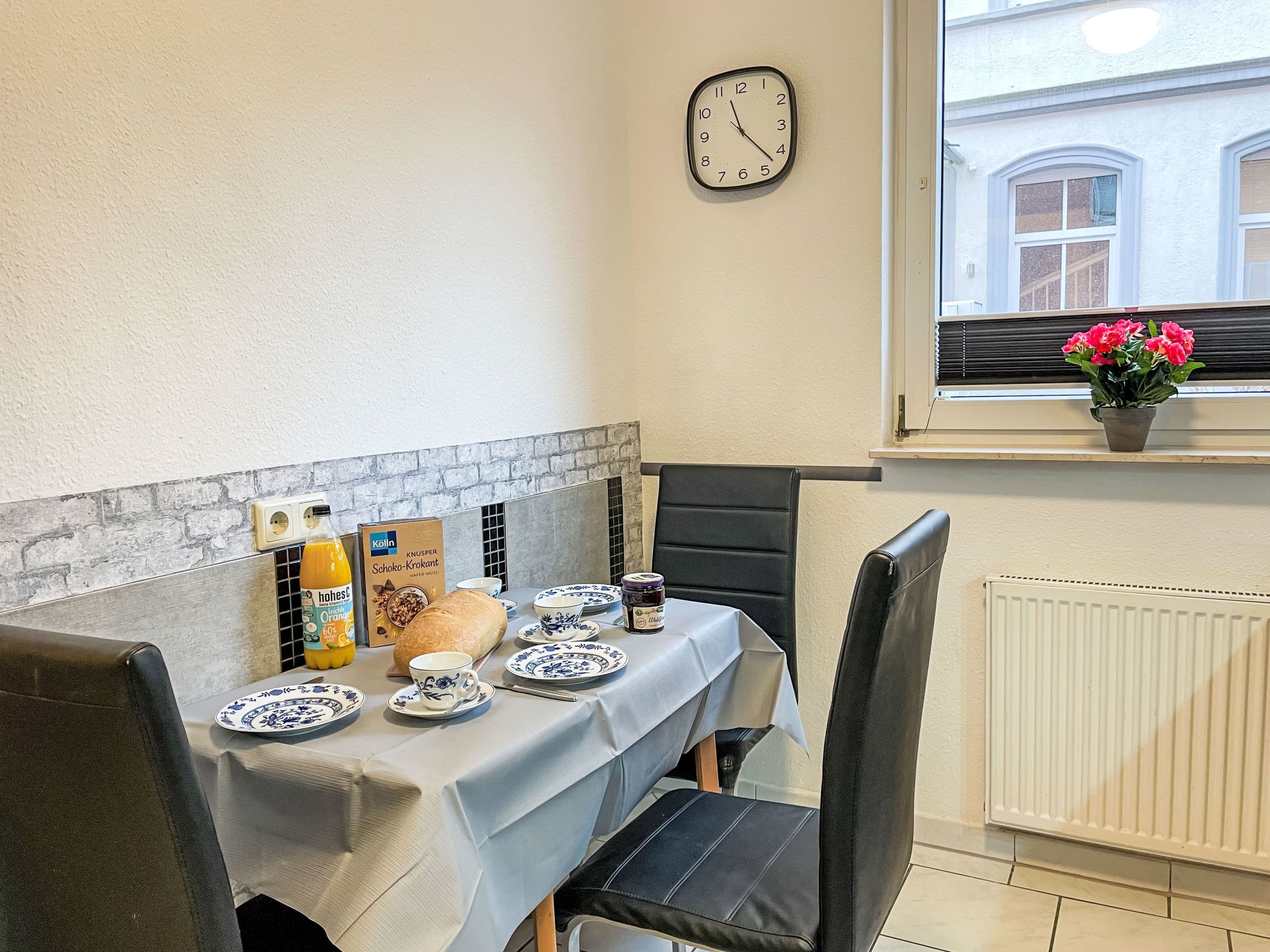 Una mesa de comedor con mantel blanco, platos, una botella de jugo de naranja y una caja de chocolate Knusper. Hay un reloj en la pared y una ventana con vistas a edificios.