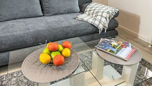 Una sala de estar con un sofá gris, una mesa de centro de vidrio con fruta y una revista, y una manta decorativa.