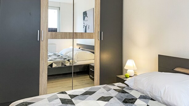 Una habitación de hotel moderna con un armario grande, una cama con cubrecama estampado geométricamente, una lámpara de noche y una mesita con una manzana.