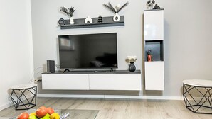 Una sala de estar moderna con un televisor de pantalla plana grande montado en una pared blanca, un mueble de entretenimiento negro debajo y una mesa de centro de vidrio con fruta.