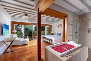 A modern bedroom with a large bed, a bathtub filled with rose petals, a shower, and a balcony with a view of the sea. - Villa Amankara (Karon)