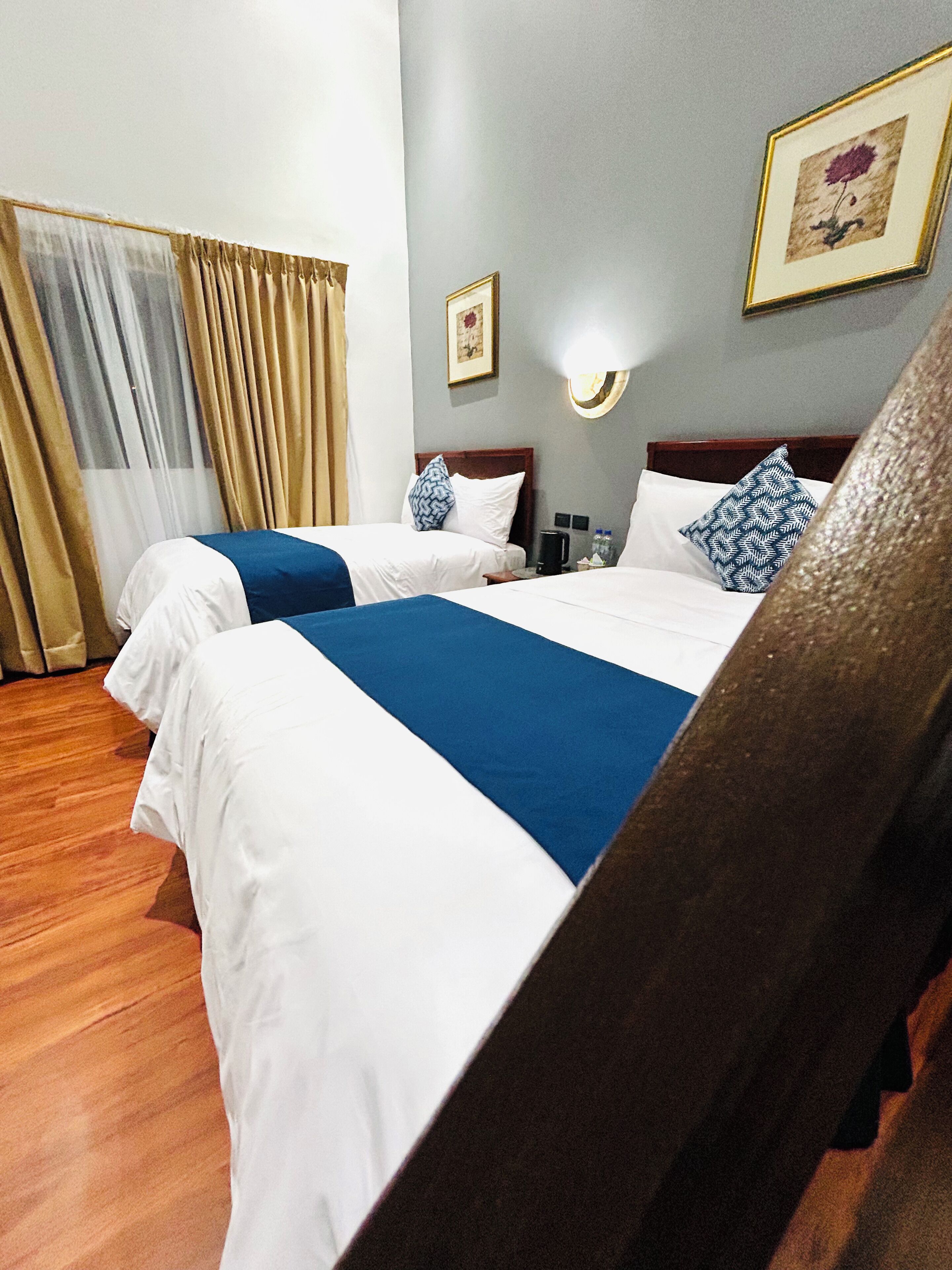 A hotel room with two beds, a wooden headboard, a nightstand, a framed picture on the wall, and a window with curtains.
