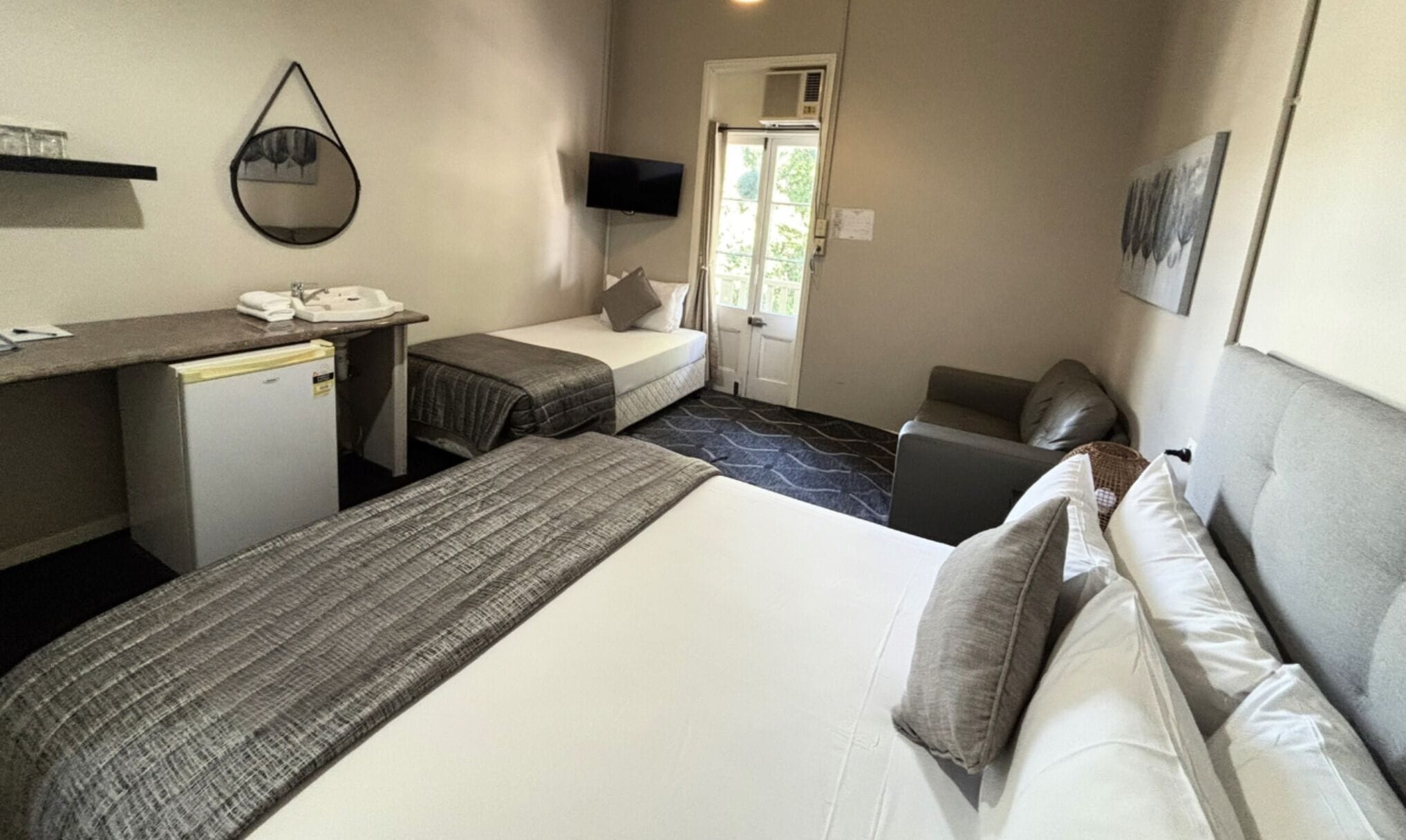 Habitación de hotel con cama, escritorio con teléfono, refrigerador pequeño, televisión y puerta que da a un balcón.