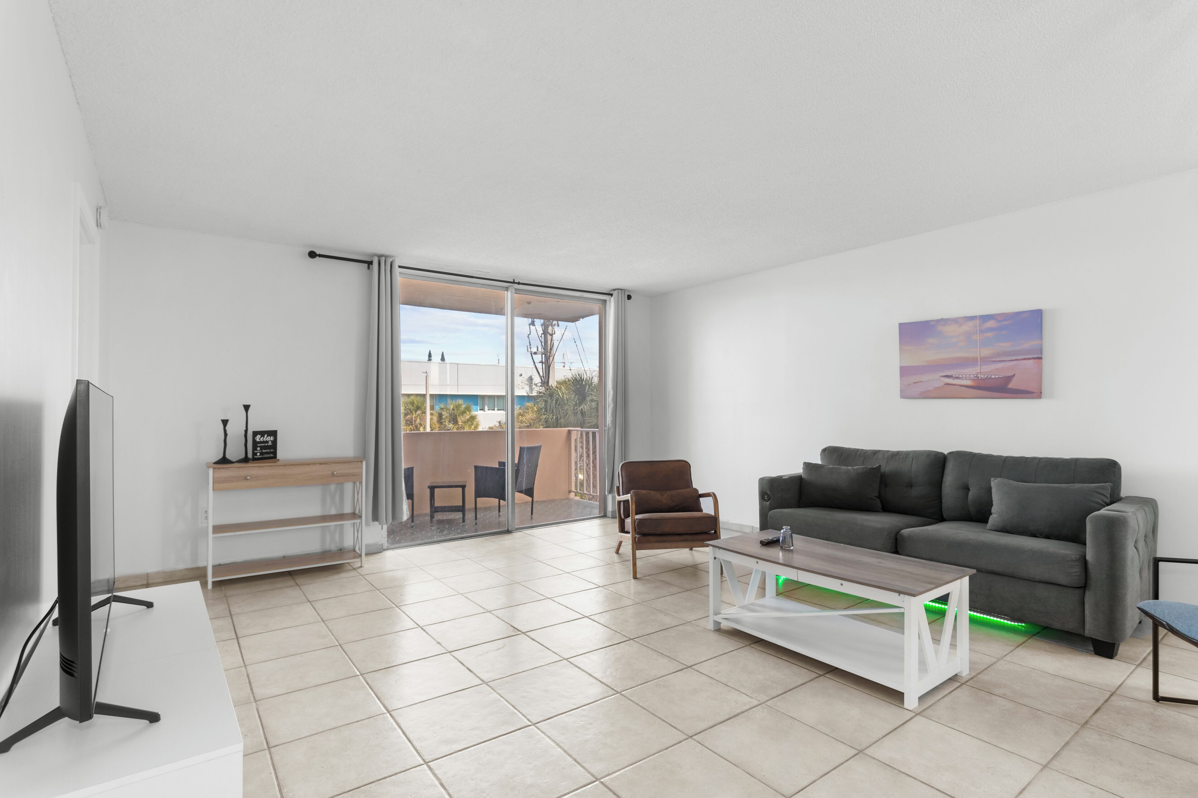 A modern living room with a flat-screen TV, a grey sofa, a brown armchair, and a glass door leading to a balcony.